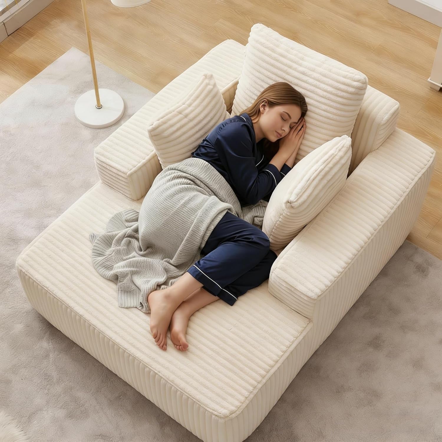 Overhead view of a cream corduroy chaise with three pillows and a woman curled up on a rug and wood floor.