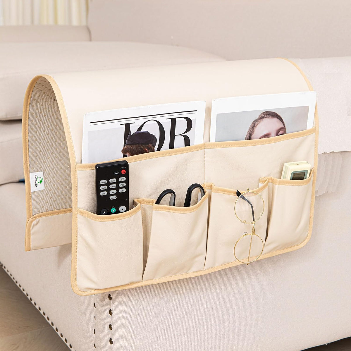Beige armrest organizer on a sofa arm showing six front pockets holding remotes, glasses and a thermometer.