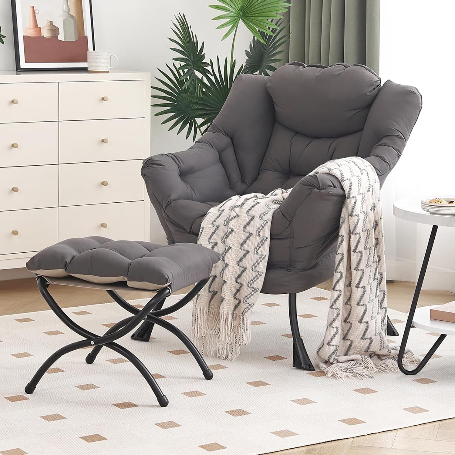 Dark grey padded lounge chair with matching ottoman, cream throw draped over arm, small side table and dresser in bright room