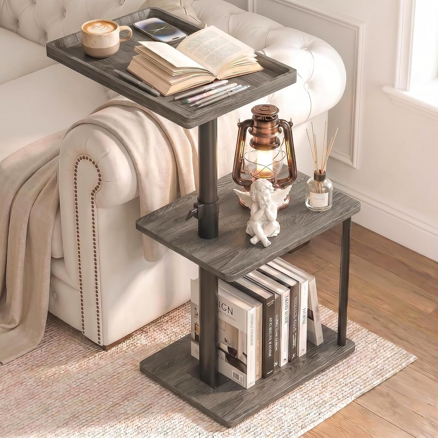 Close detail of a gray wood three-tier C‑shaped table beside a sofa with an open book, pens, a glowing lantern and stacked