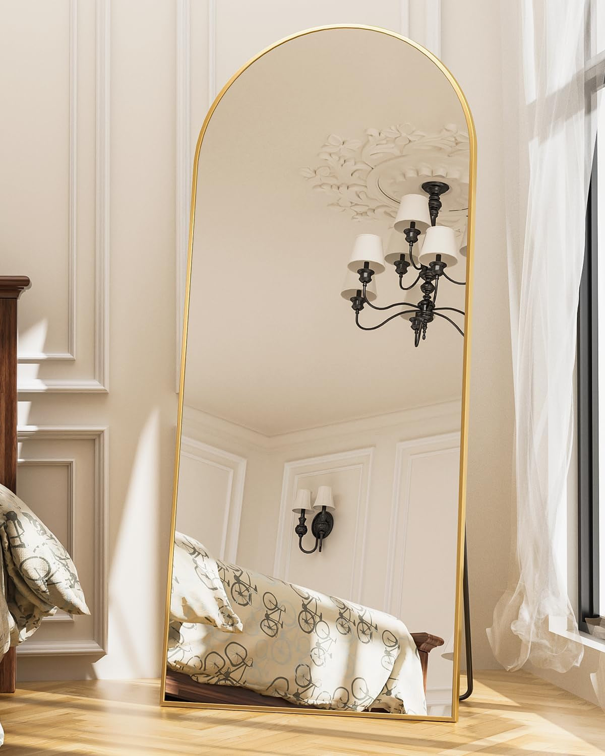 Arched full-length mirror with slim gold frame leaning against wall, reflecting chandelier and bed
