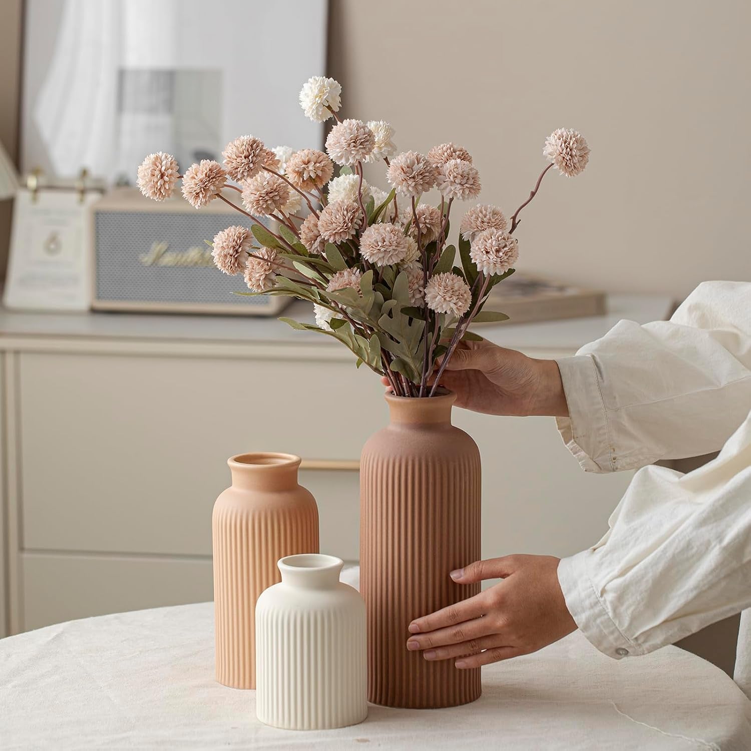 Hand placing faux flowers into the tall terracotta ribbed vase with two smaller matching vases on a dining table.