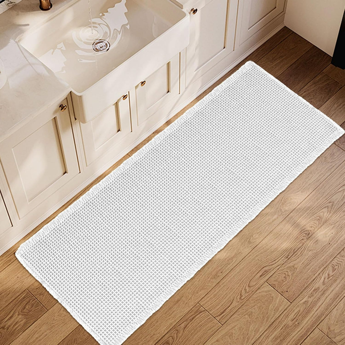 Top-down view of a white waffle-texture bath mat placed in front of a farmhouse sink on wood floor.