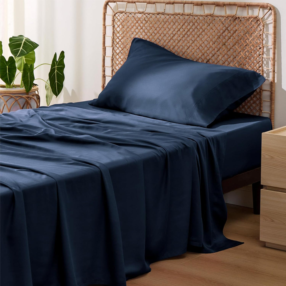 Three-quarter shot of a bed with deep navy sheets and pillow, drape of fabric pooling onto a wood floor.