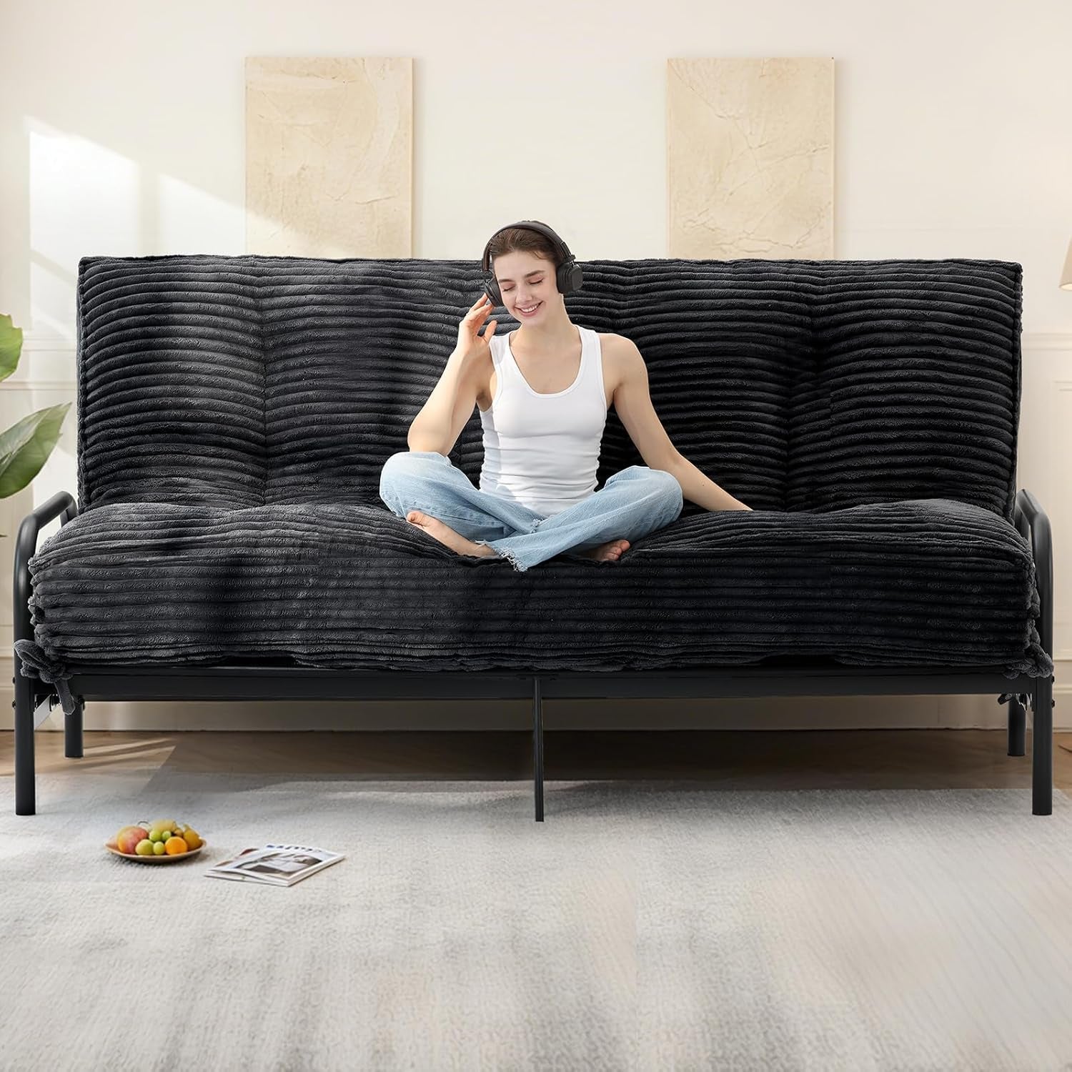 Woman wearing headphones sitting cross-legged on a dark gray ribbed faux-fur futon mattress on a black metal frame in a