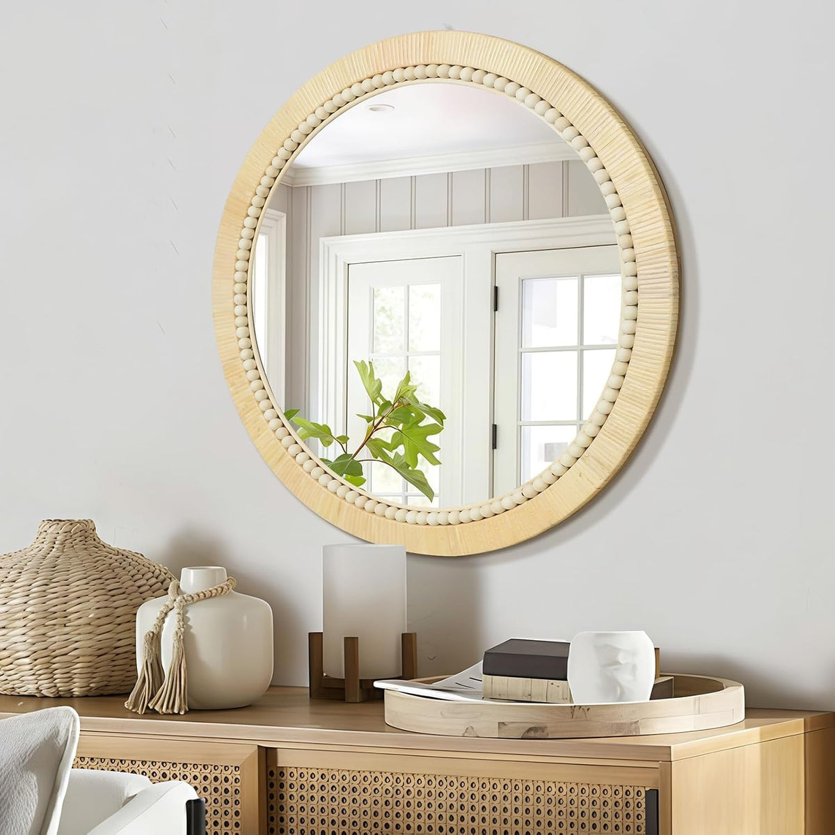 Angled side view of a round woven rattan mirror with bead accent hung above a wooden console decorated with vases and books