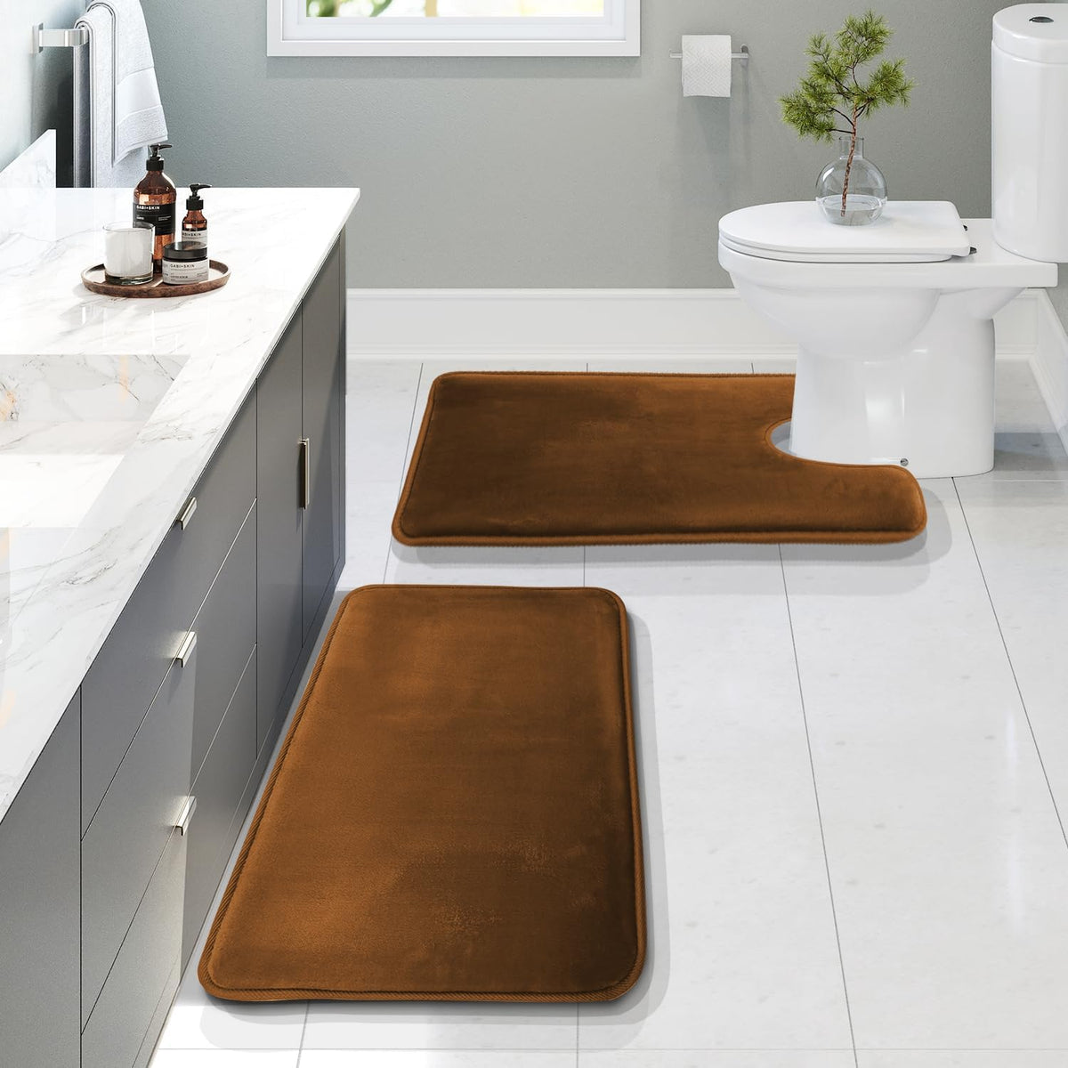 Brown memory-foam contour rug placed at the toilet with a matching brown rectangular rug along the vanity on white tile
