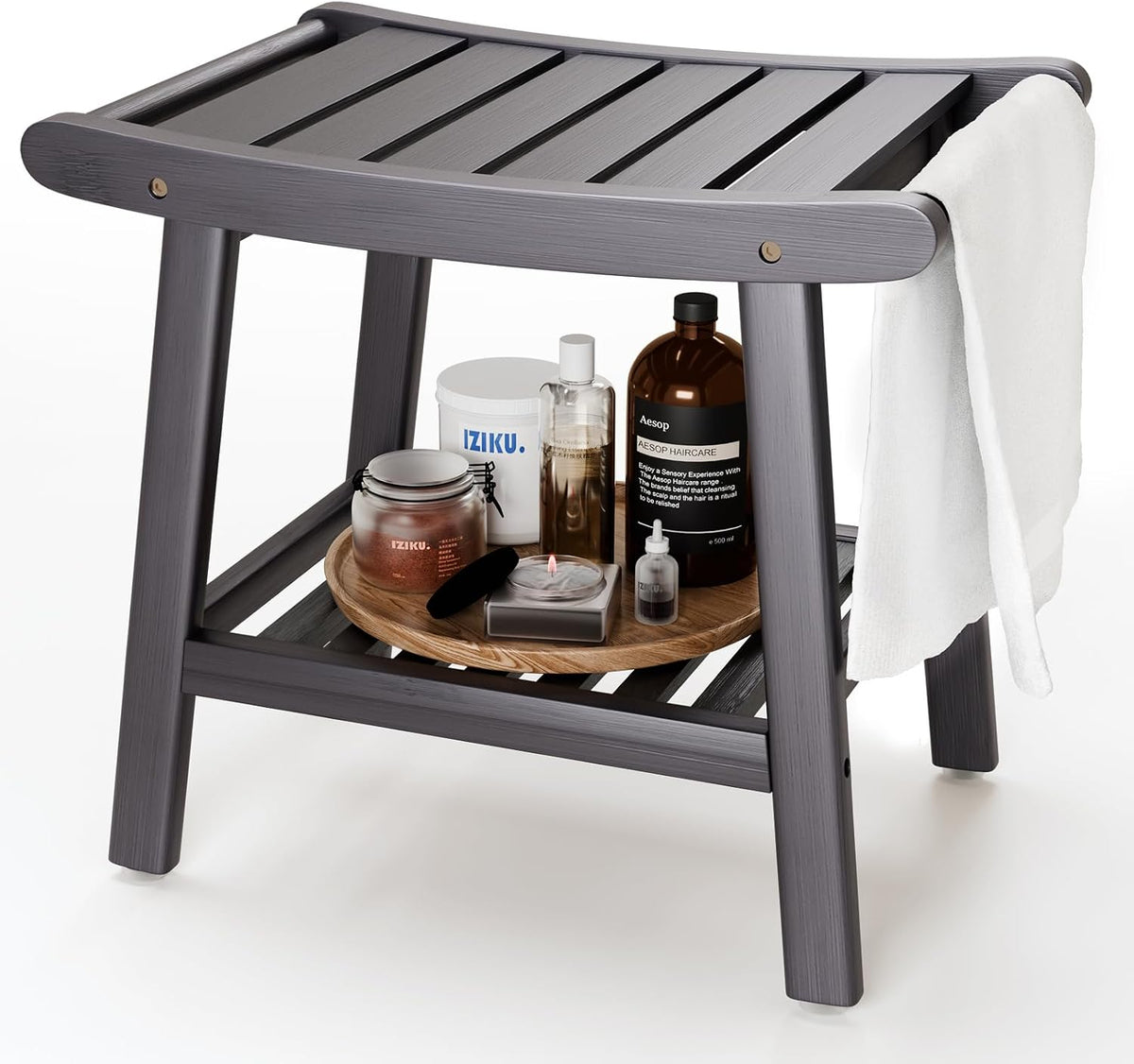 Angled view of a gray slatted bamboo shower stool with lower shelf holding bath bottles and a towel draped on the side.