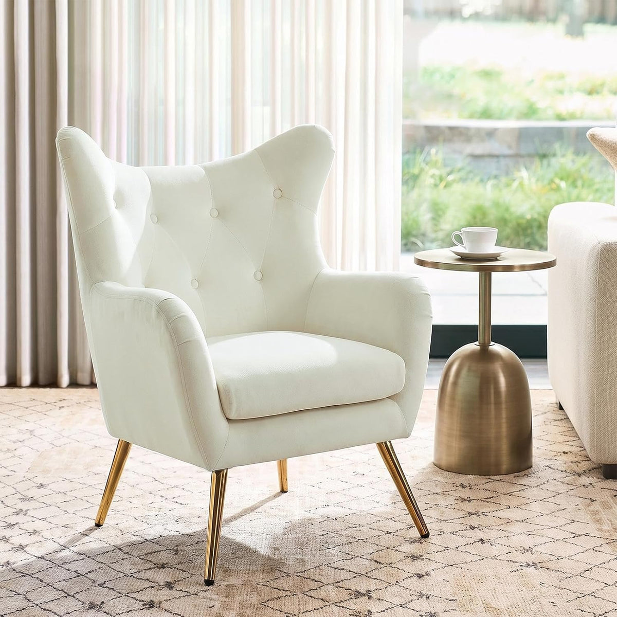 Off-white wingback chair shown at a three-quarter side angle with a slim gold side table and sunlit window.
