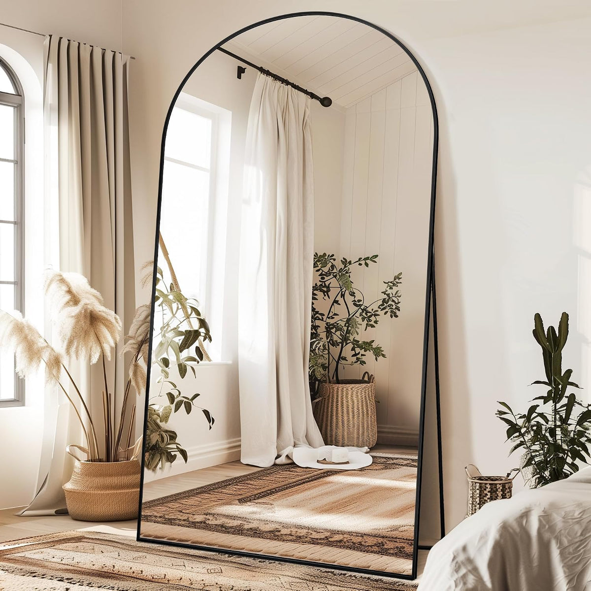 Tall arched mirror with slim black frame standing on a rug near windows and potted plants.