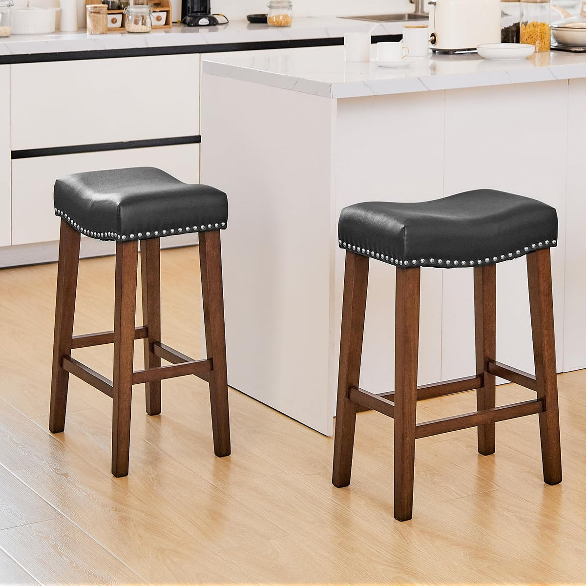 Two backless black saddle stools with nailhead trim at a white kitchen island on light wood floor.
