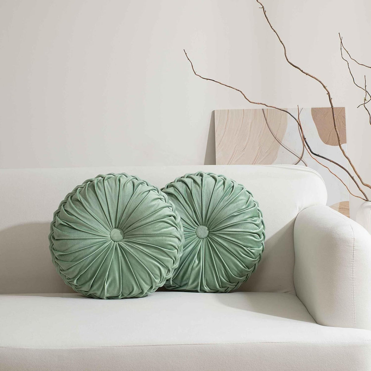 Pair of round ruched pillows in soft sage green arranged on a light sofa armrest