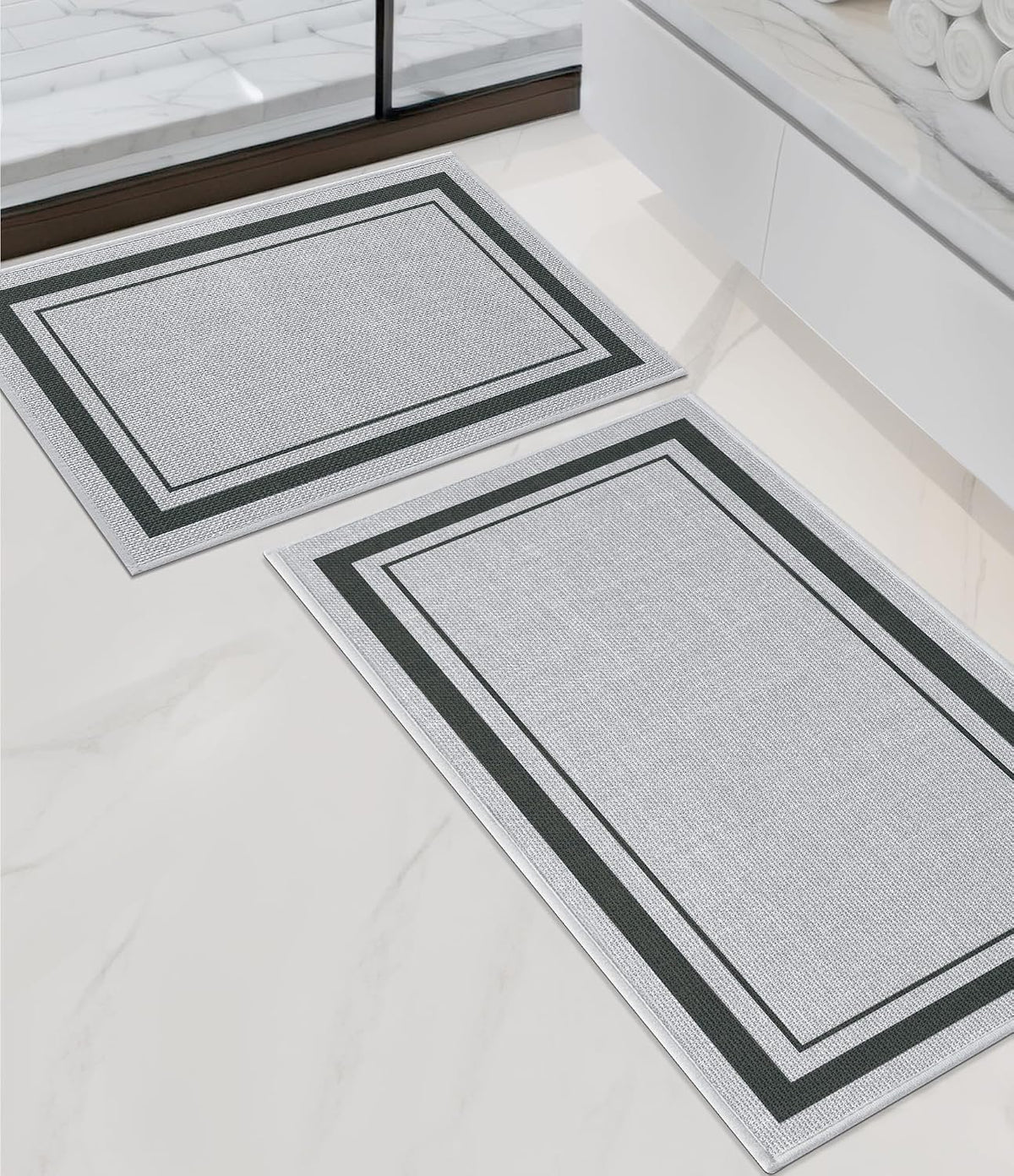 Angled bathroom view of two light-gray rectangular rugs with double dark-green borders beside a white vanity.
