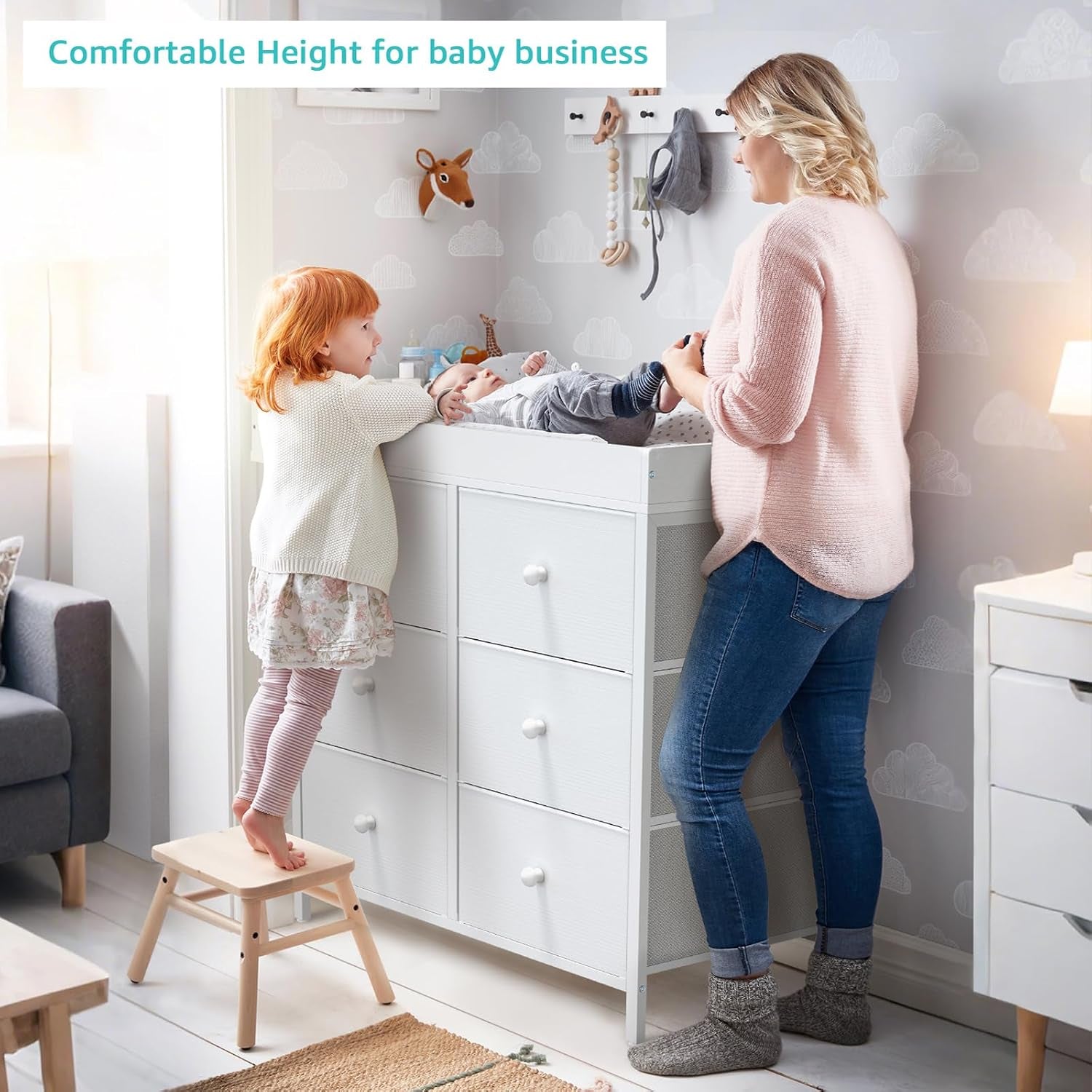 Woman and young child using the white changing dresser in a softly lit nursery with wallpaper and a stool.