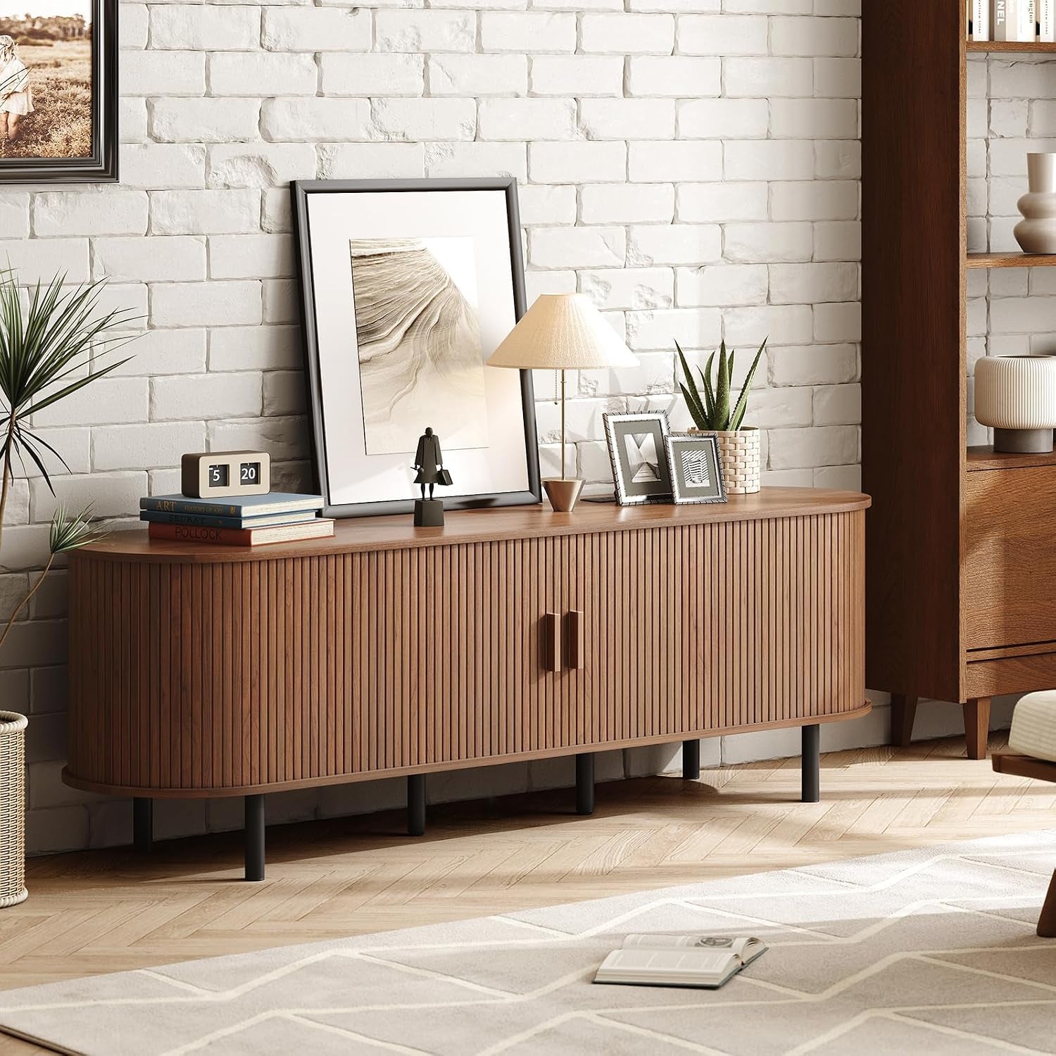 Low-angle living room view of the walnut stand against a white brick wall with lamp, framed art and plants on top.