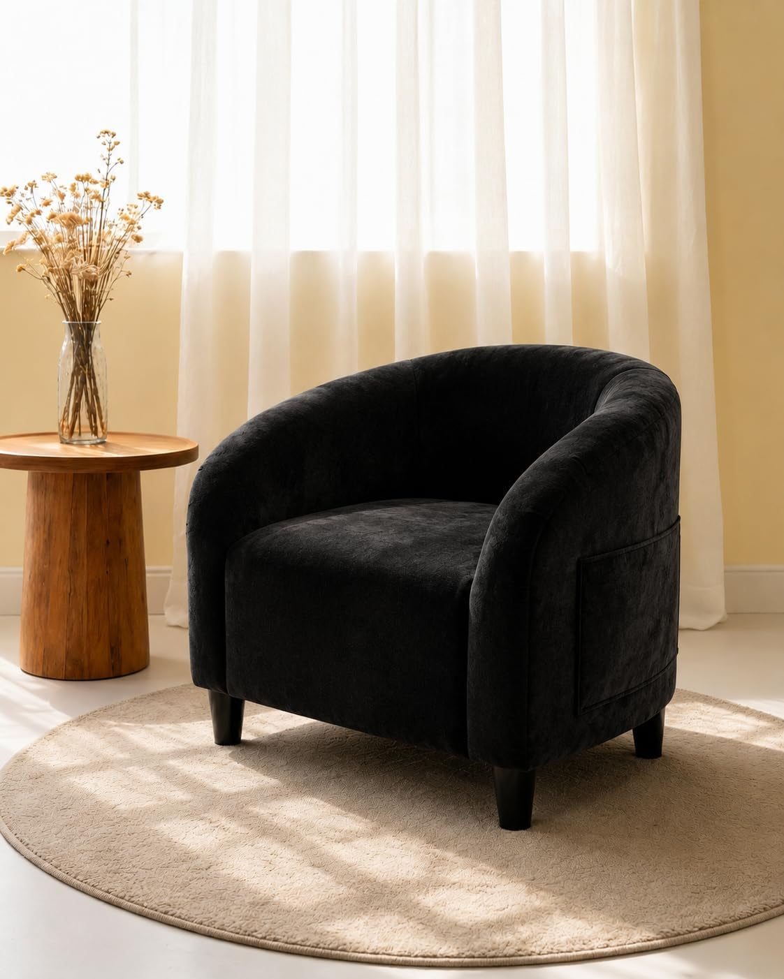 Black barrel armchair on a circular rug next to a wooden side table and vase under a sheer window.