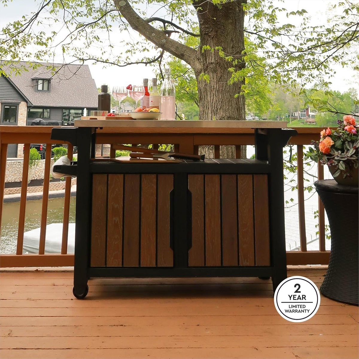 Full view of the outdoor storage cabinet on a deck with stainless top, bottles and glasses on top and a lake beyond
