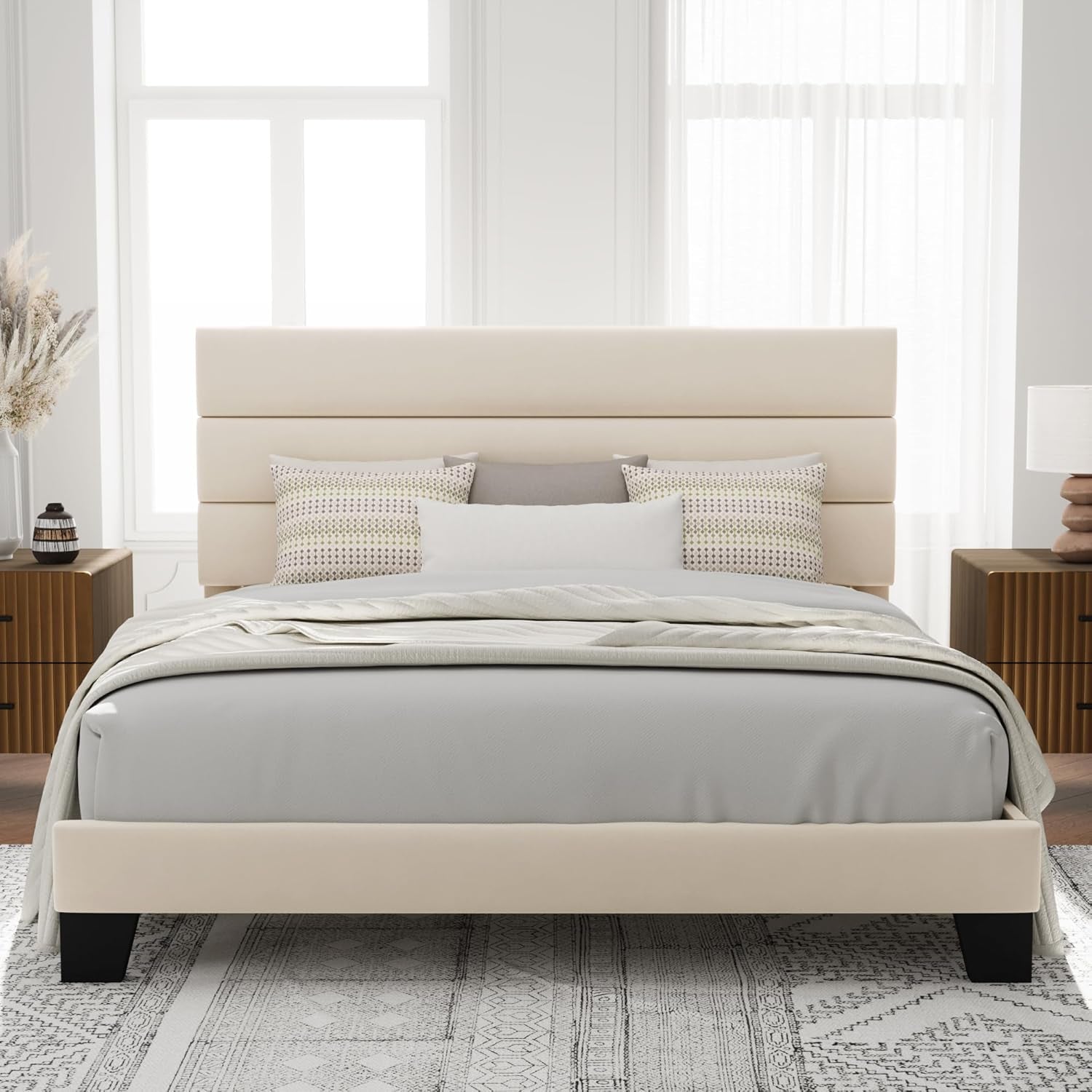 Front view of a beige upholstered bed with a three-panel headboard and matching footboard in a bright bedroom.