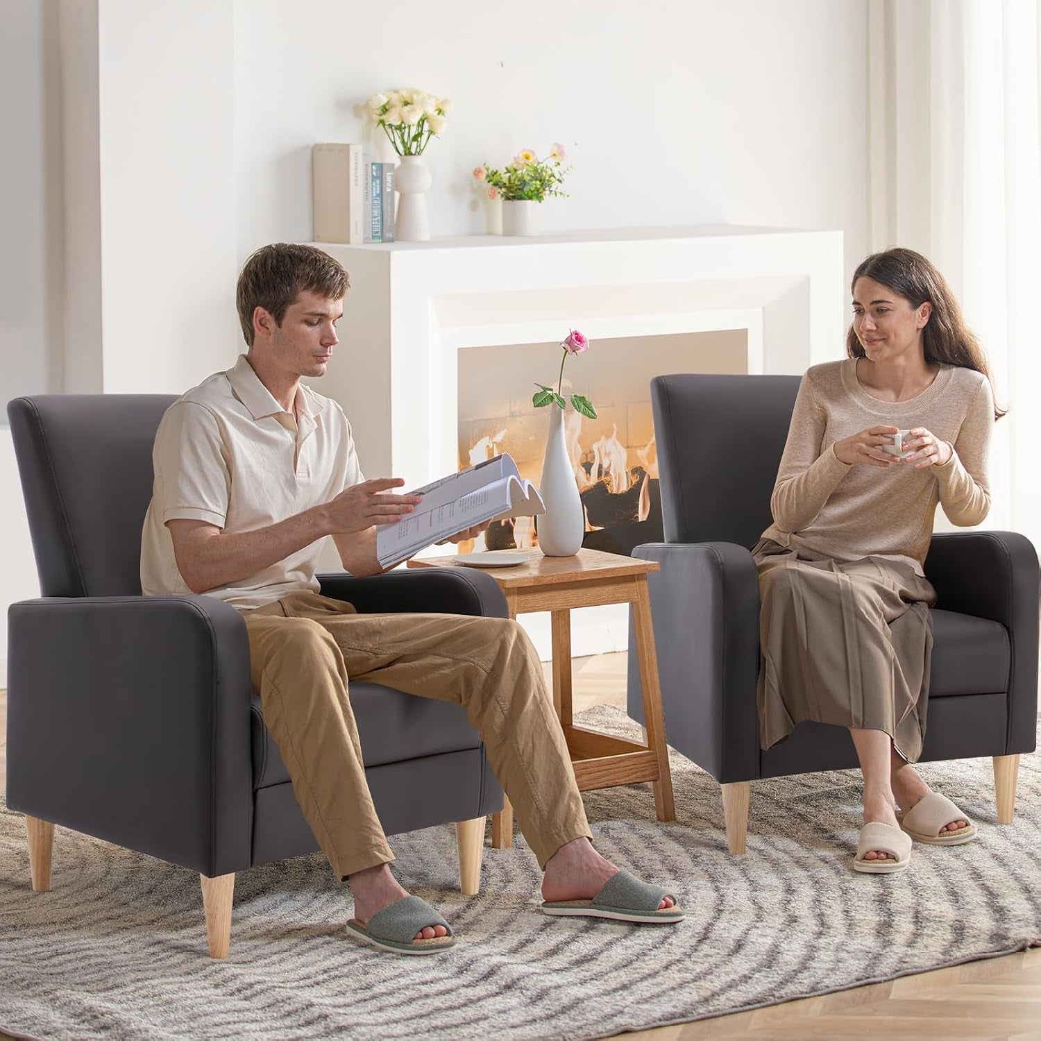 Two people seated in matching high-back chairs with wood legs beside a small table and fireplace