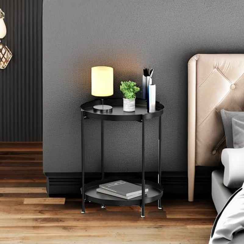 Black two-tier round metal side table next to a bed holding a small lamp, potted plant, and pens.