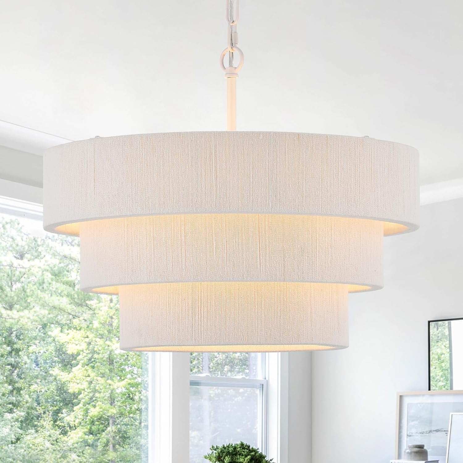 Lit three-tier white drum pendant hanging over a bright room with windows and wall art