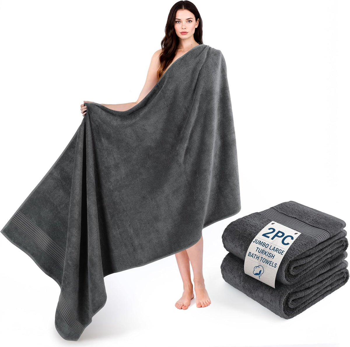 Woman holding a jumbo charcoal-gray towel with a matching folded stack of towels beside her on a white background.
