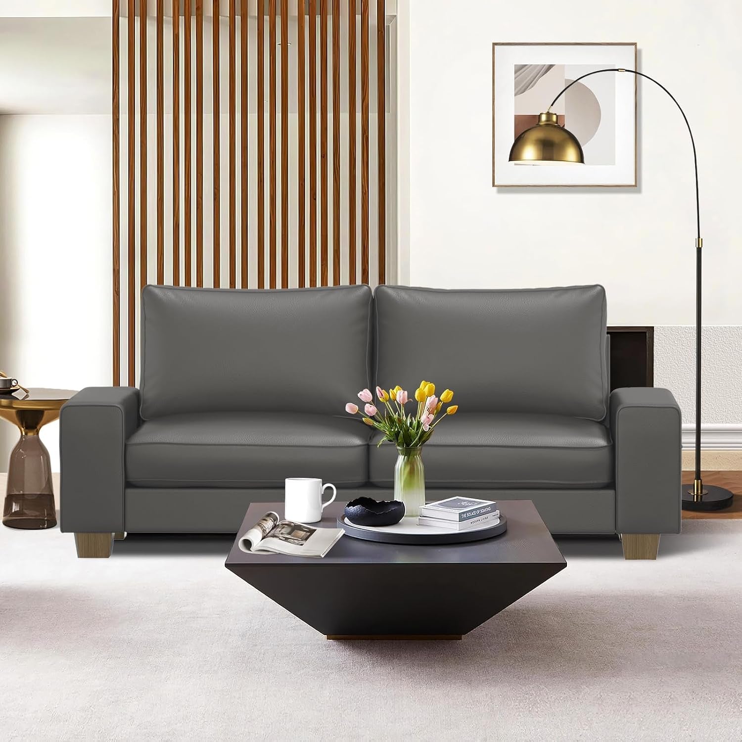 Front view of a dark gray faux-leather loveseat in a staged living room with a low geometric coffee table and floor lamp