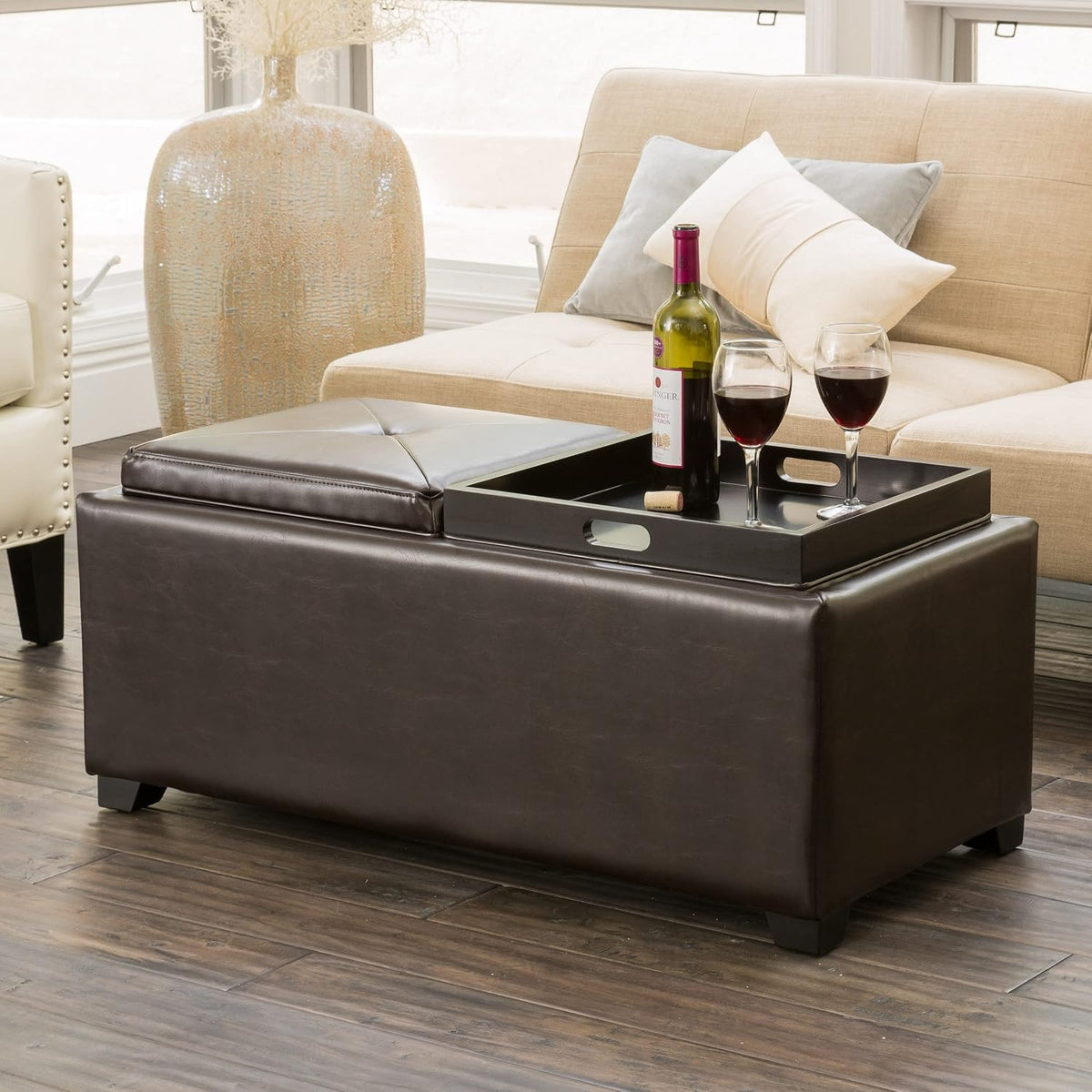 Brown ottoman in a living room with a black tray holding a bottle of wine and two filled wine glasses.