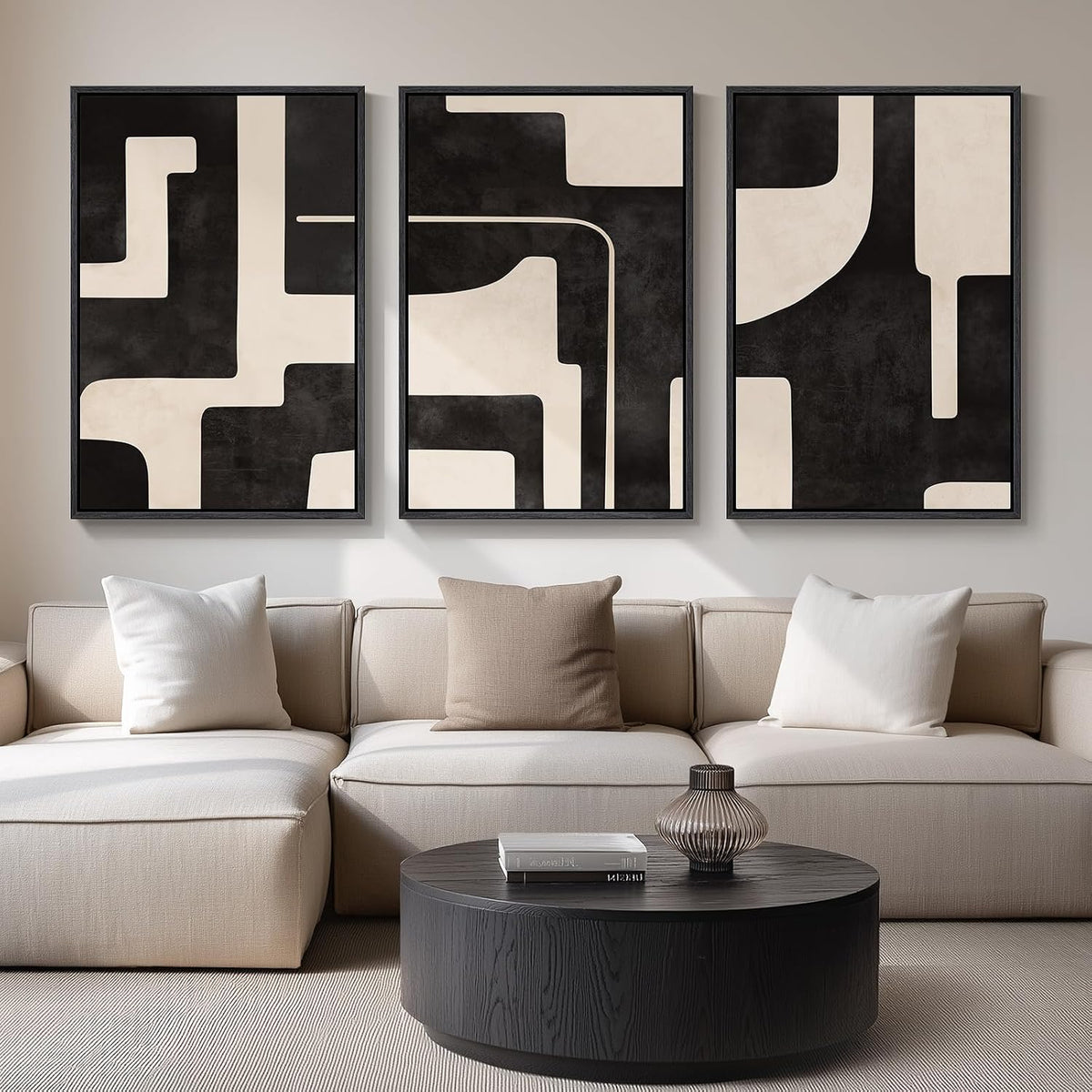 Three framed abstract panels with large black and cream geometric shapes above a neutral sofa and round dark wood table.