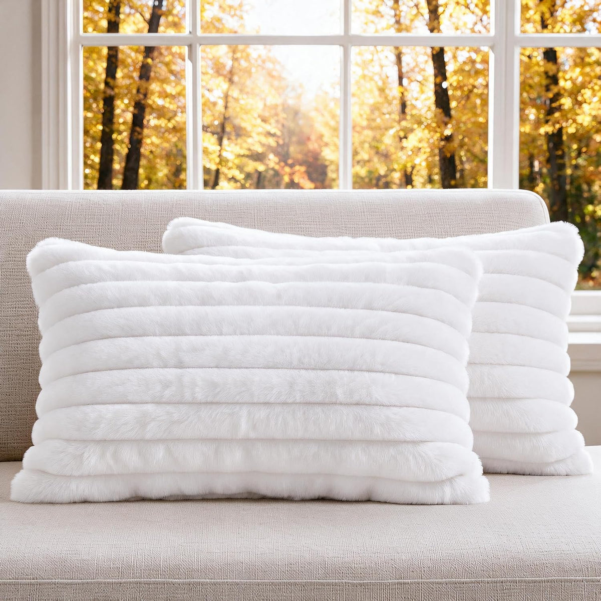 Pair of white ribbed faux-fur square pillow covers arranged on a beige sofa with fall foliage outside.