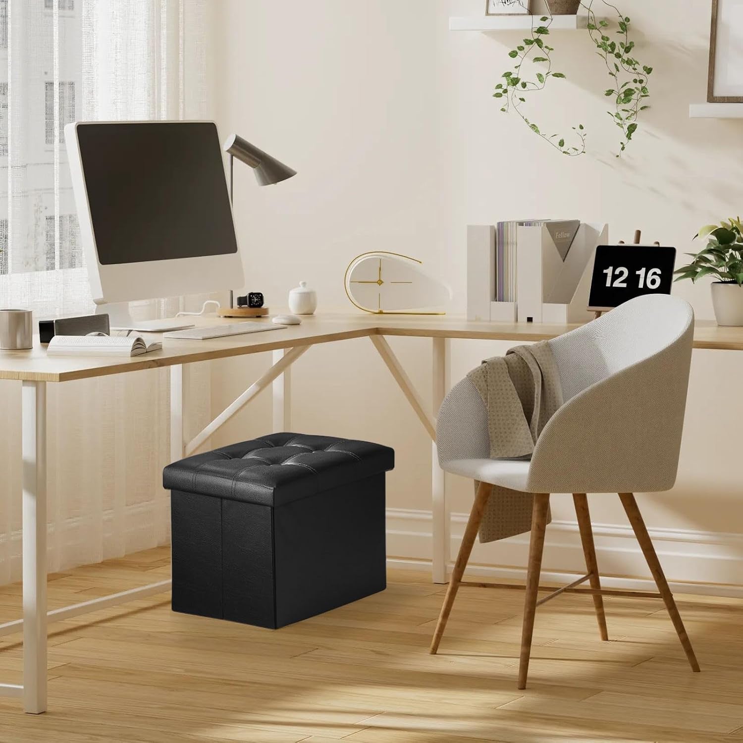 Black tufted storage ottoman placed under a desk in a bright home office next to a grey upholstered chair