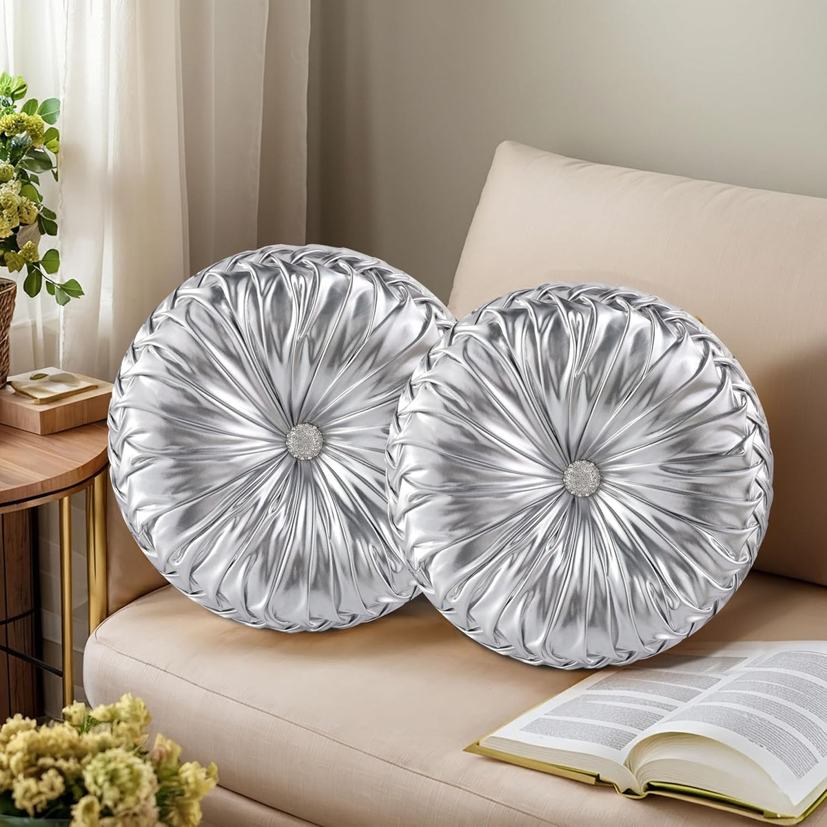 Two round silver ruched pillows with jeweled centers on a beige sofa beside an open book.