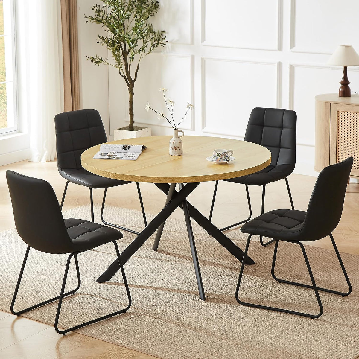 Round light-wood dining table with four black U-shaped sled chairs in a bright room.
