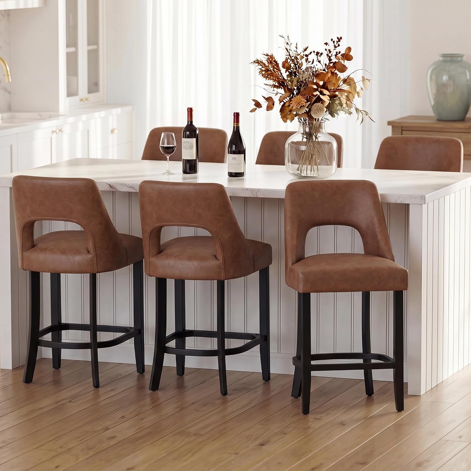 Three brown upholstered counter stools lined at a white paneled island with a vase and wine