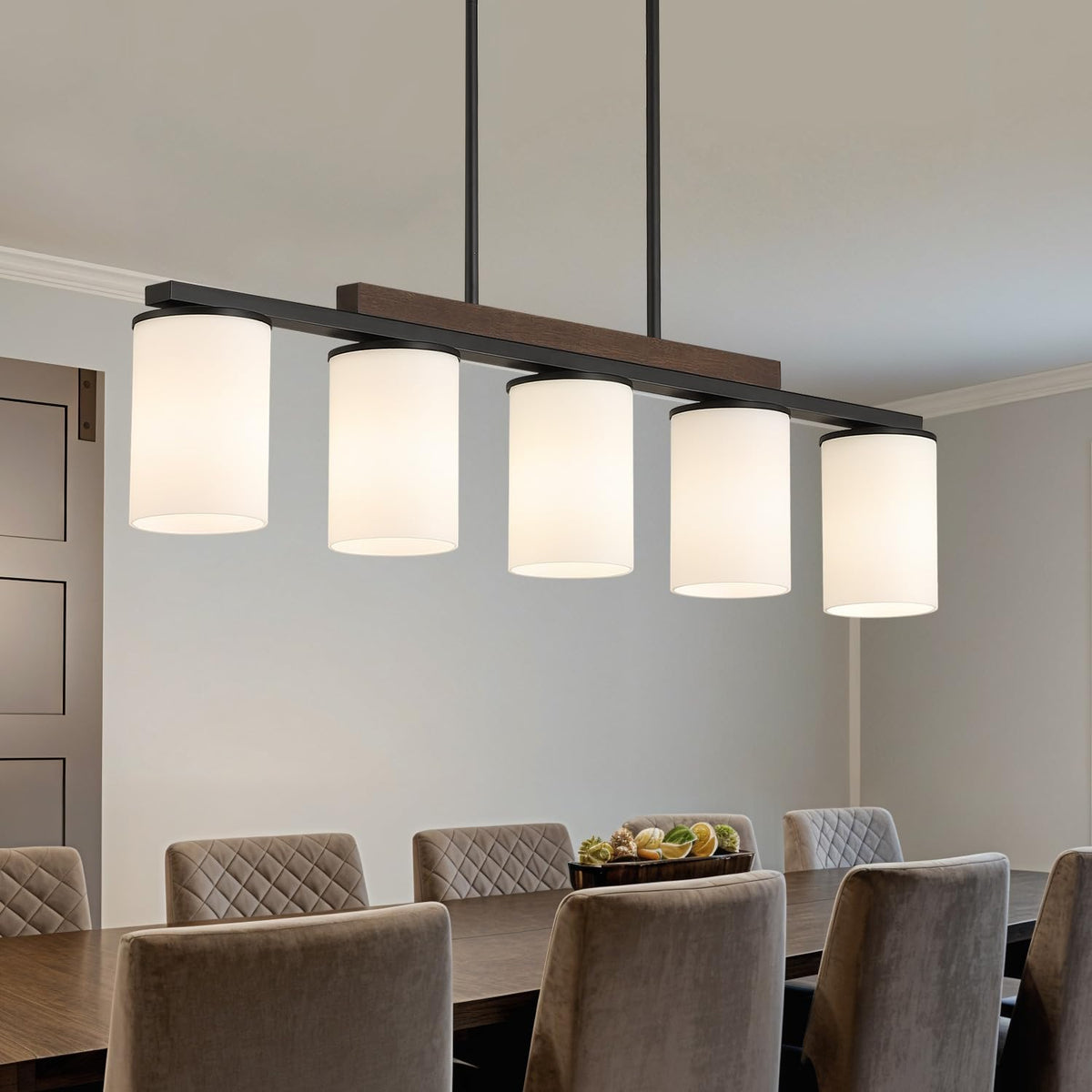 Side view of a five-light linear chandelier with white drum shades suspended over a long dining table.