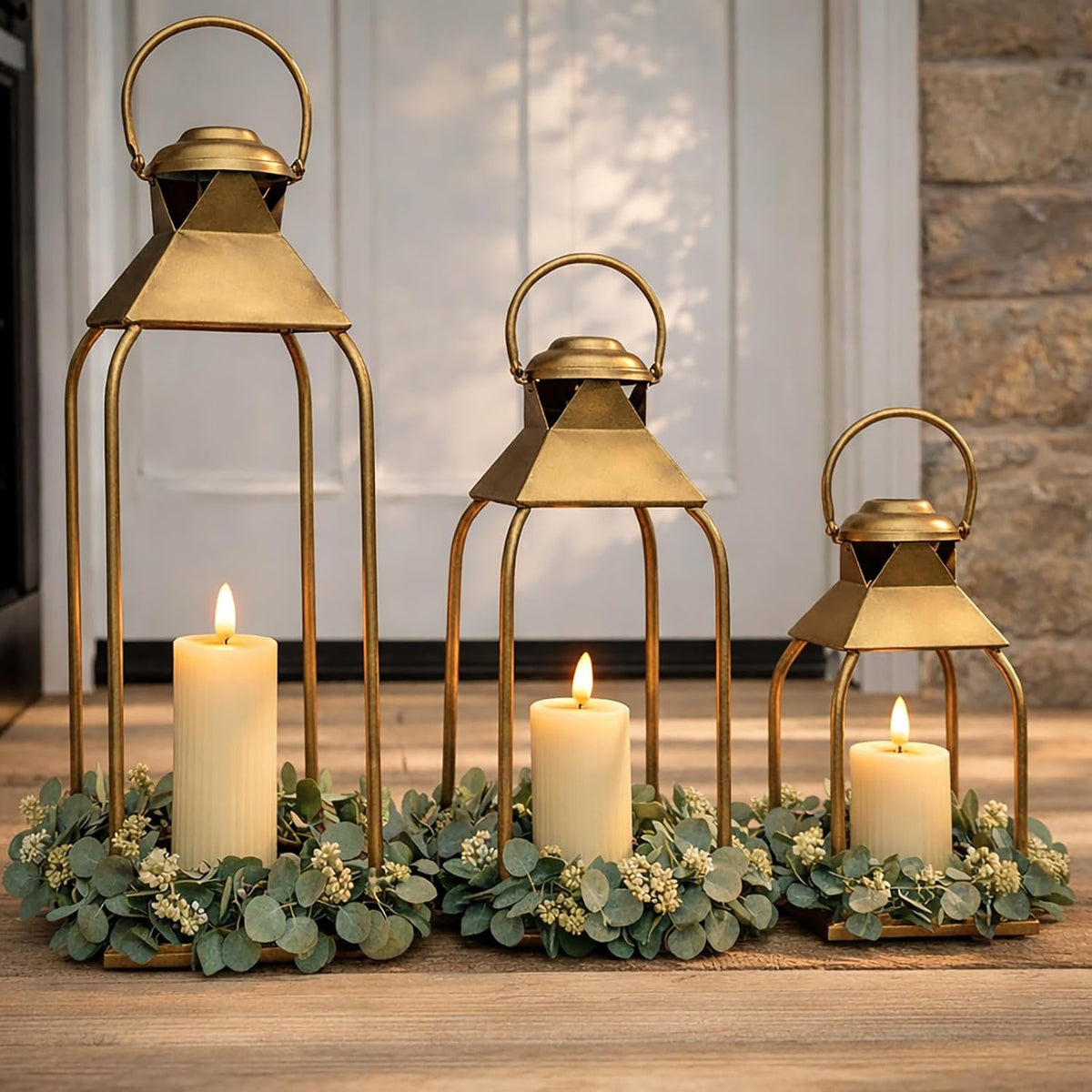 Three antique gold lanterns of different heights arranged on a table with candles and greenery in front of a door.