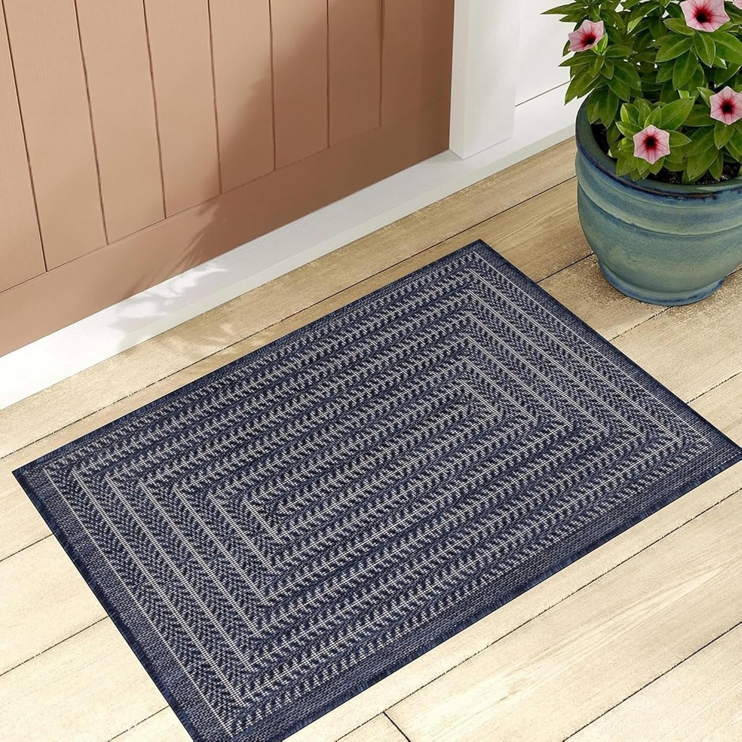 Navy rectangular outdoor rug placed on light wood porch near a potted flowering plant.