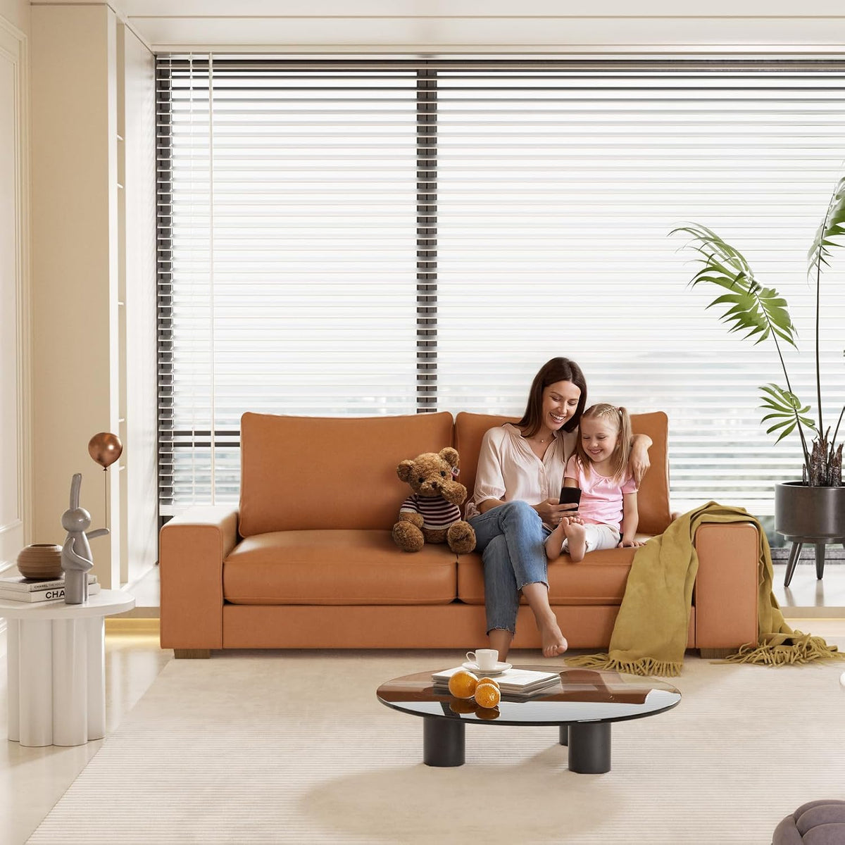 Front view of a caramel faux-leather loveseat with a woman and child sitting together by large horizontal blinds