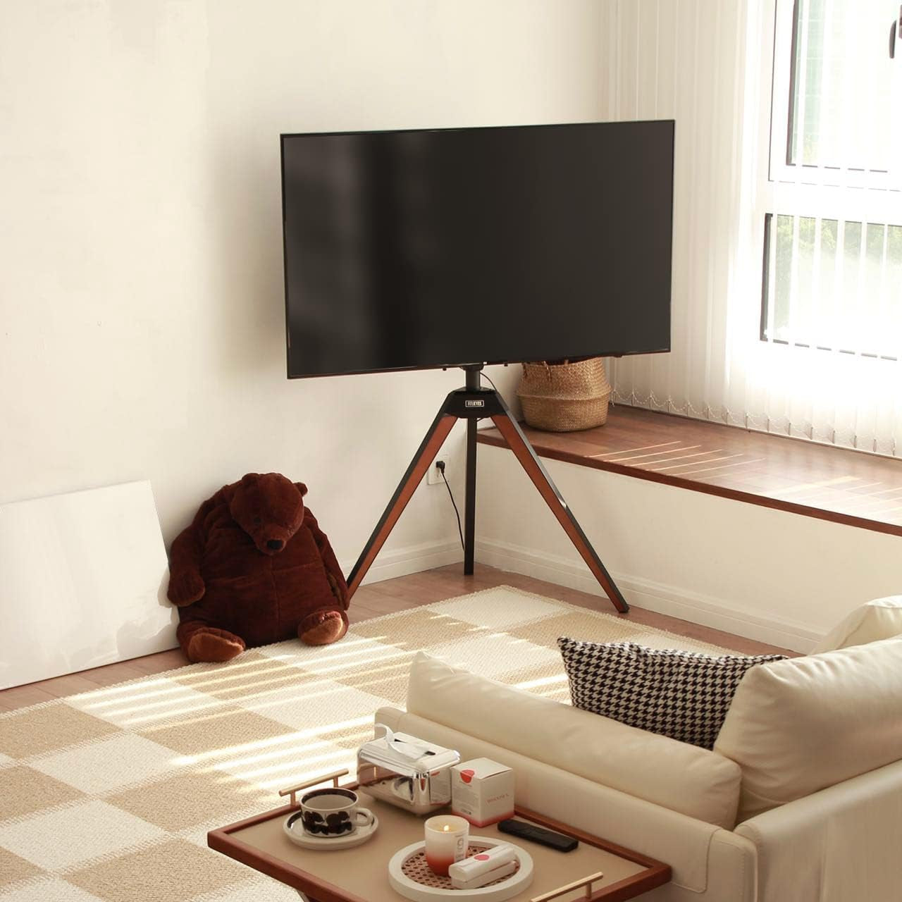 Tripod TV stand placed in a bright corner beside a window in a minimal living room setting.