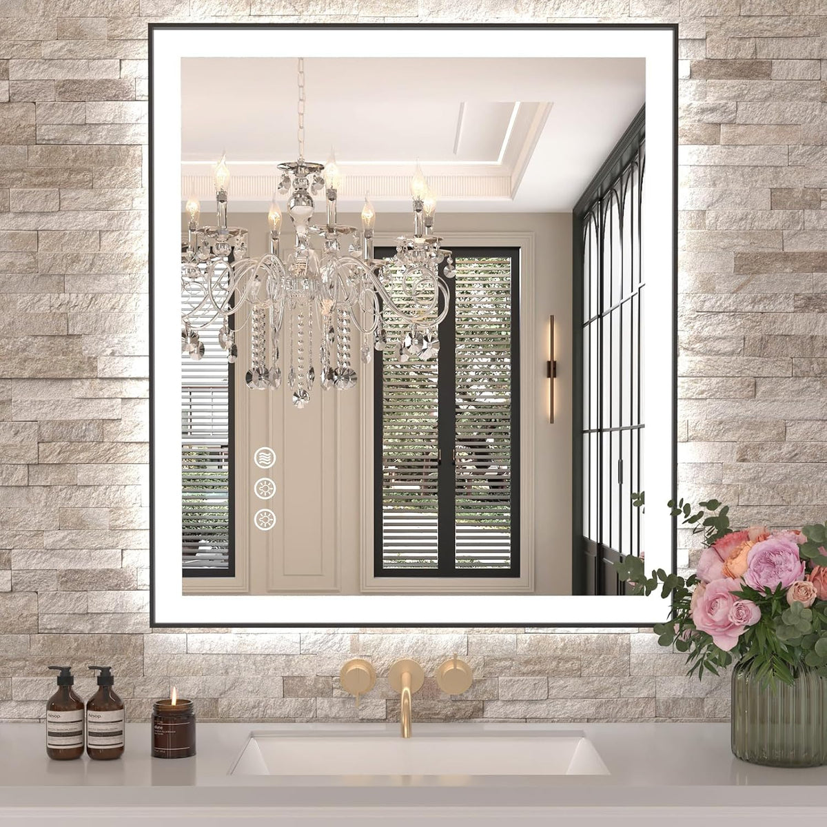 Rectangular LED mirror with glowing border above a sink, reflecting a crystal chandelier; stone wall and a vase of pink