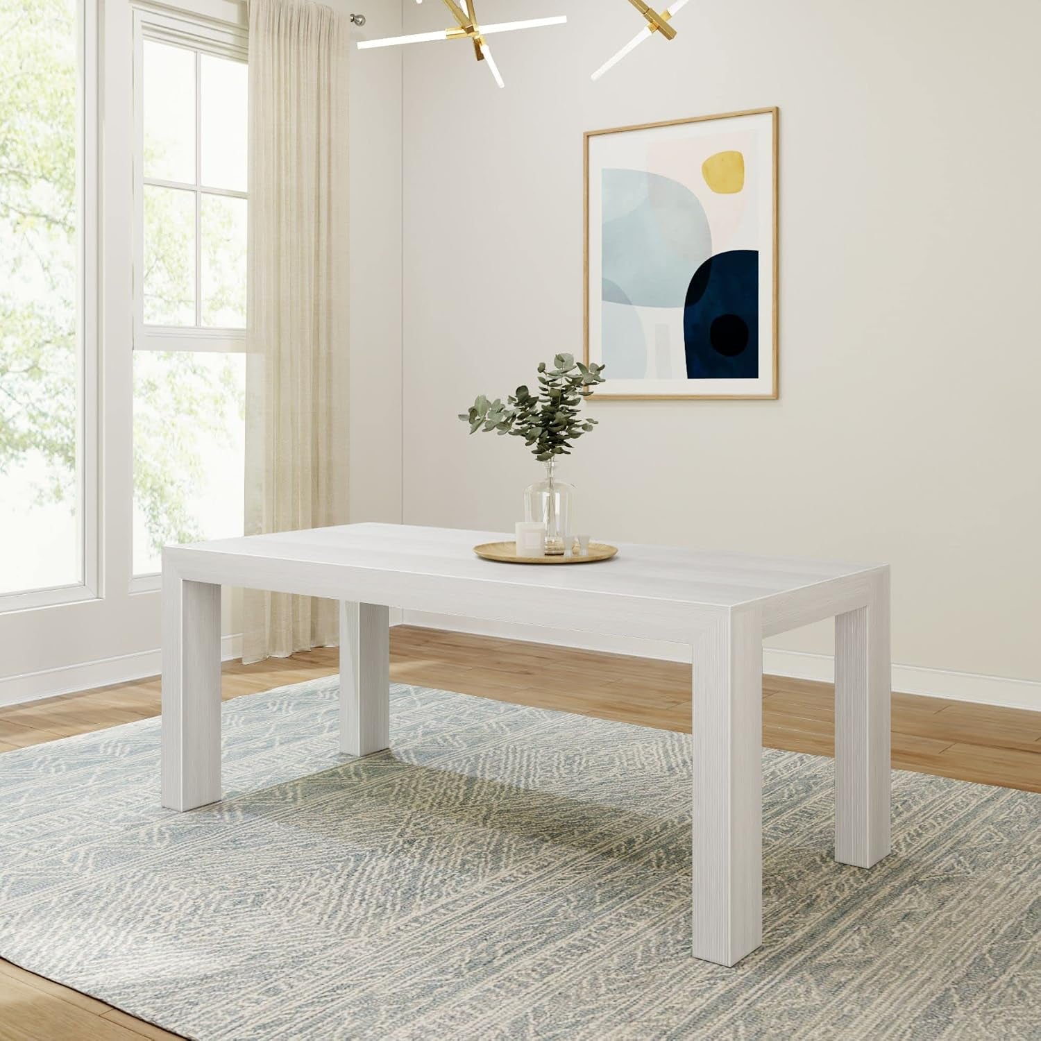 Rectangular whitewashed dining table on a patterned rug with vase and tray centerpiece.