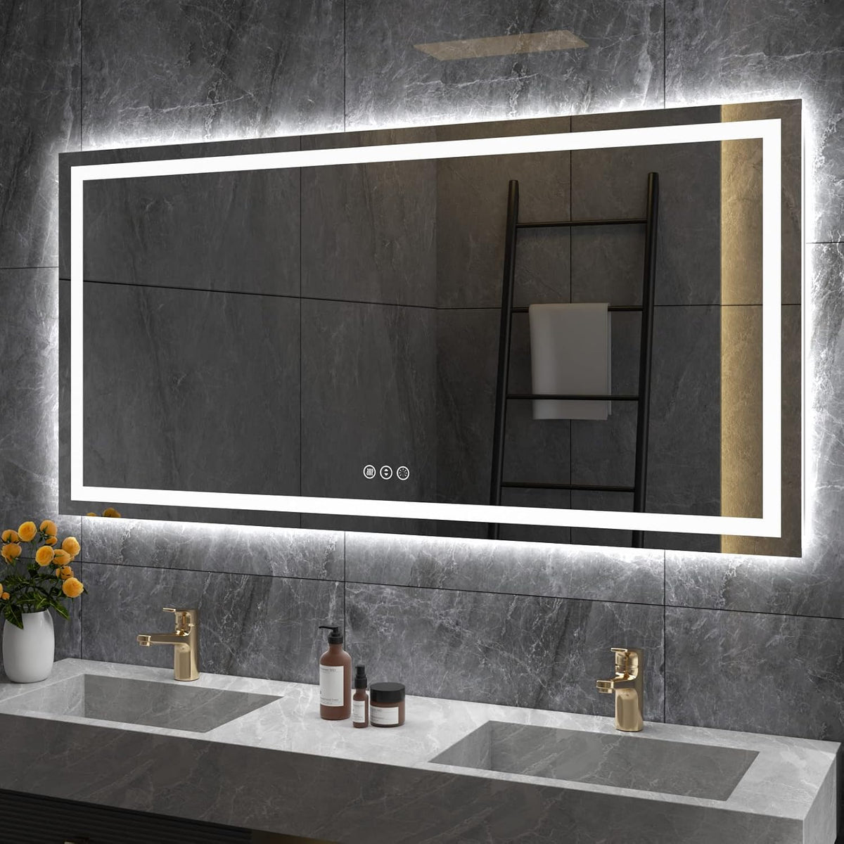 Rectangular backlit mirror mounted on dark gray stone tiles above two undermount sinks and gold faucets.