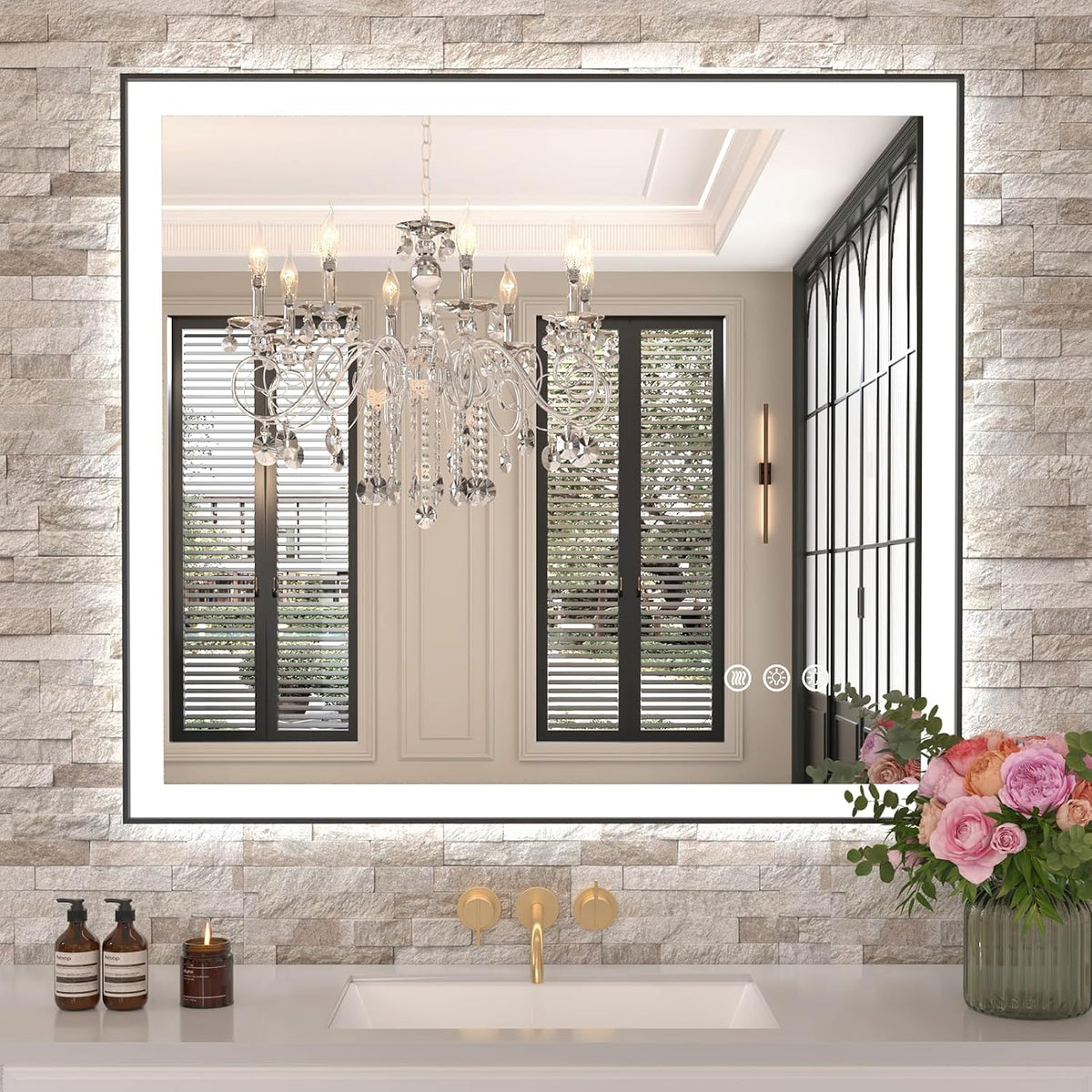 Square illuminated mirror on a light stone wall reflecting a chandelier, with a gold faucet and bouquet at the sink.