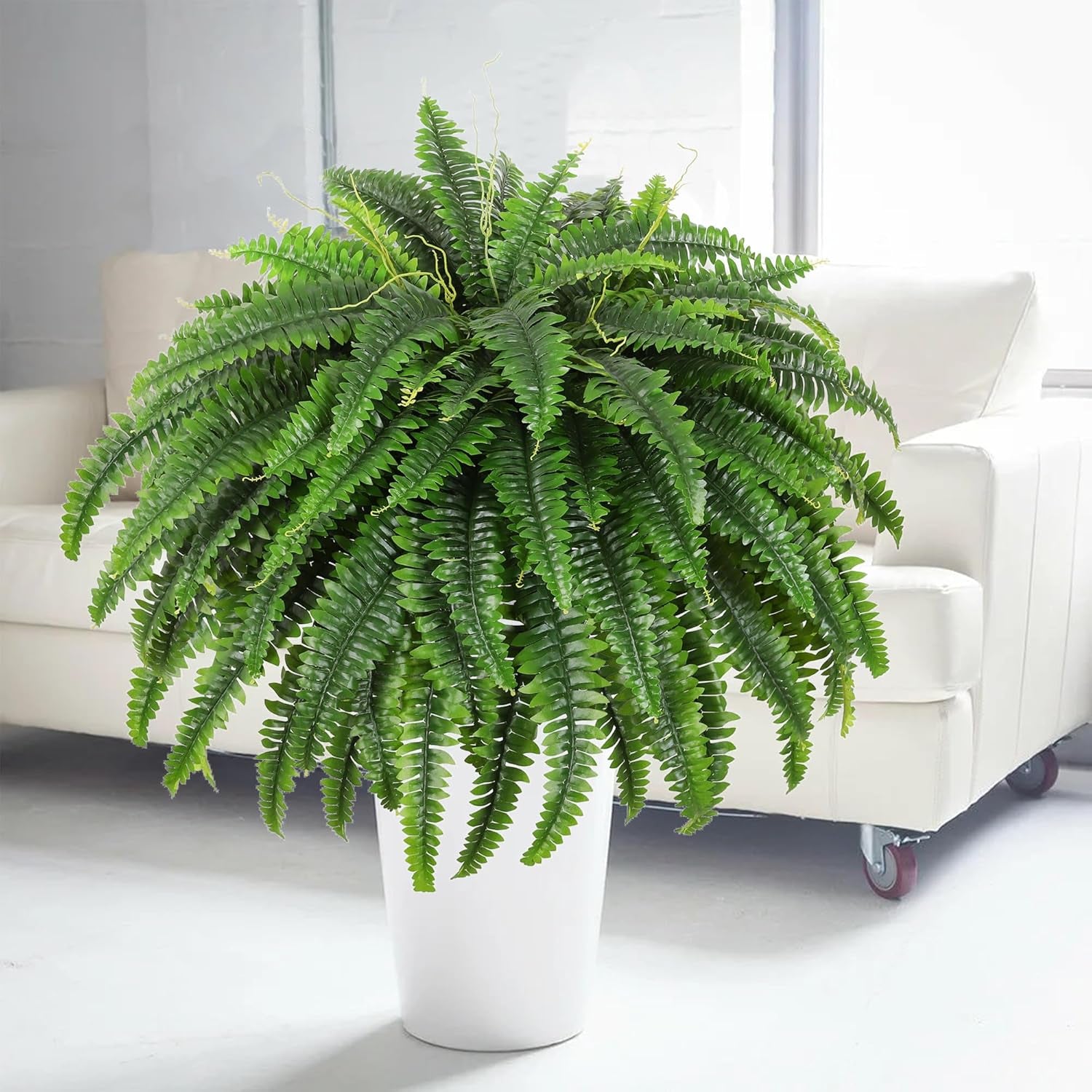 Green artificial Boston fern bush in a white cylindrical planter beside a white sofa