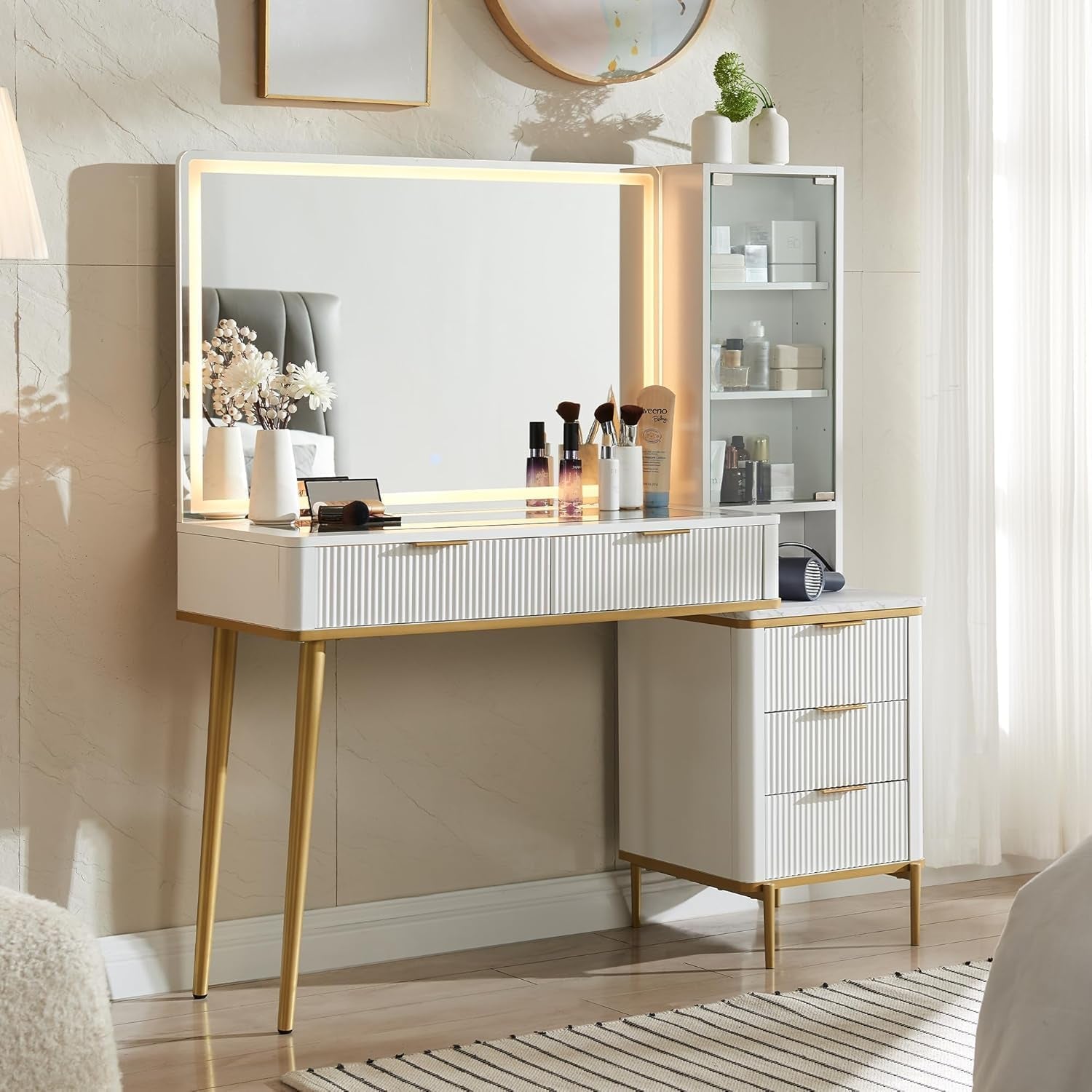 White fluted vanity with LED-framed mirror, gold trim and pedestal drawers next to a glass display cabinet