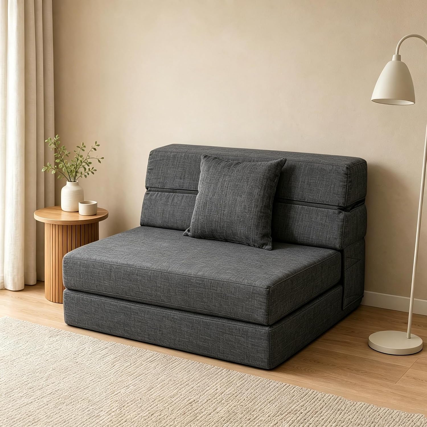 Front-left view of dark grey futon sofa bed set against a neutral living room with side table and lamp