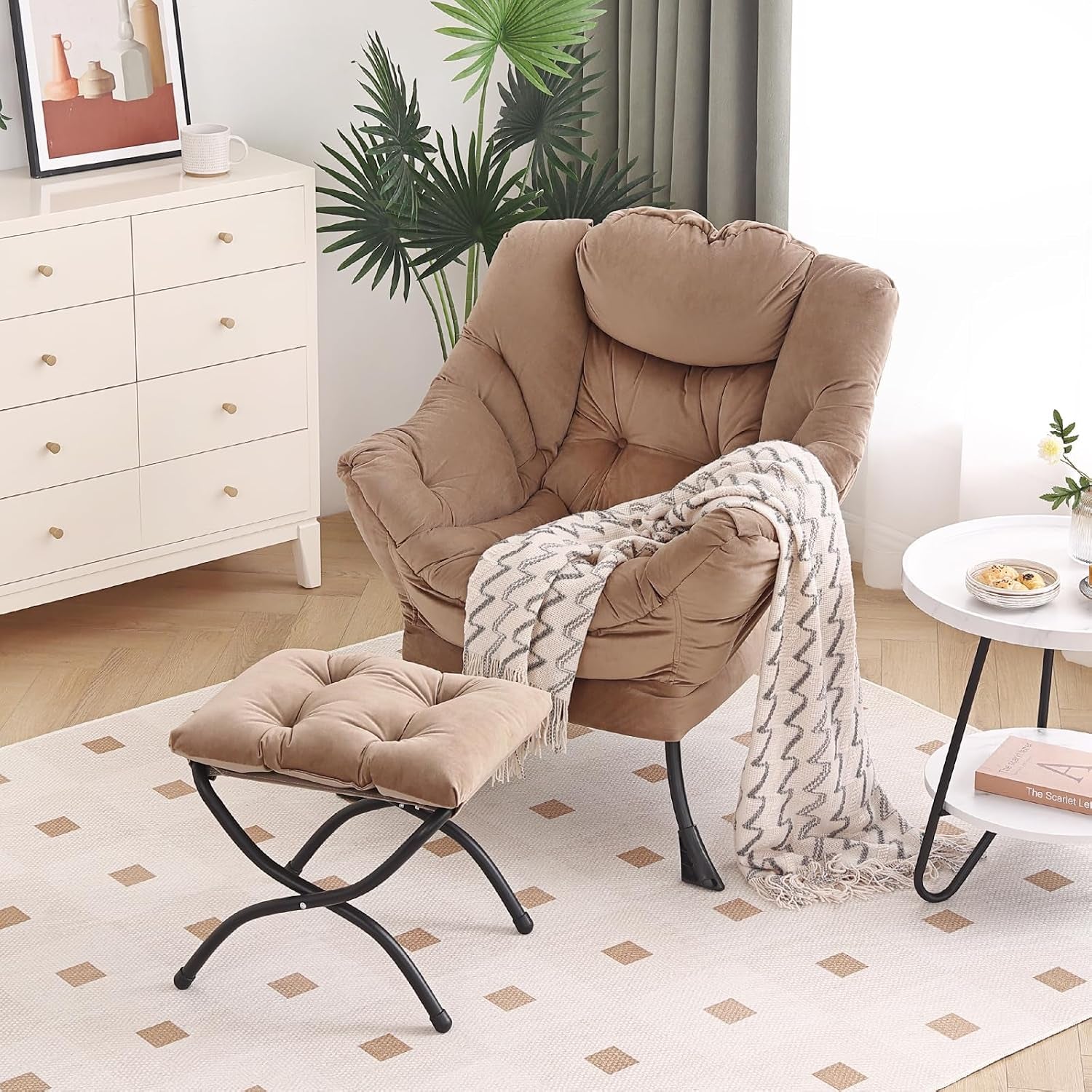 Beige cushioned lounge chair with ottoman and draped throw in a styled living space with dresser and round side table.