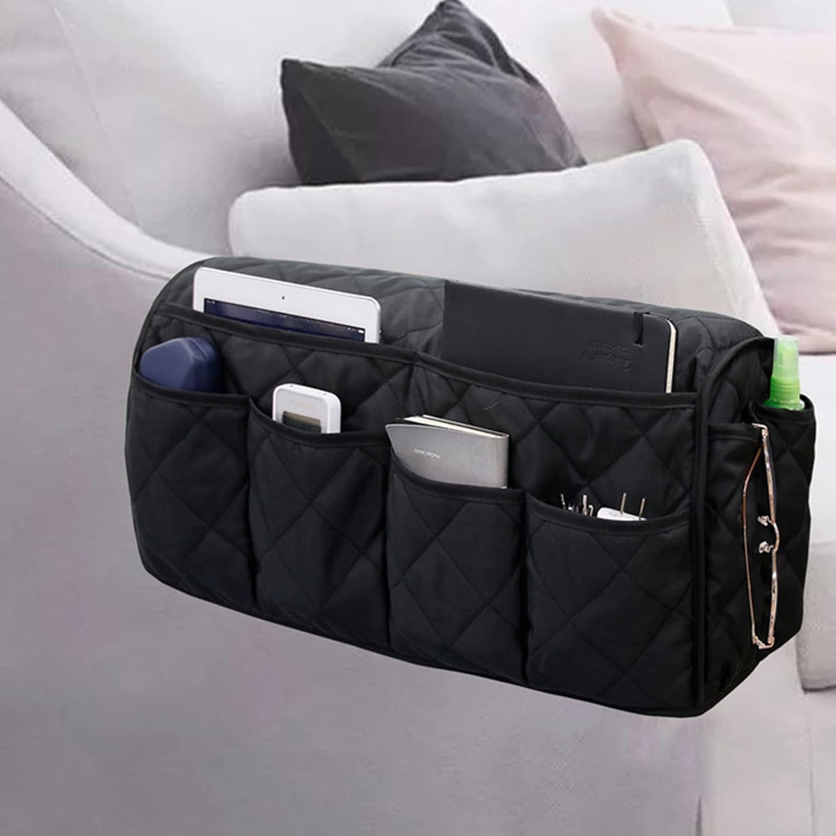 Black quilted armrest organizer draped over a sofa arm with multiple pockets holding a tablet, phone and glasses