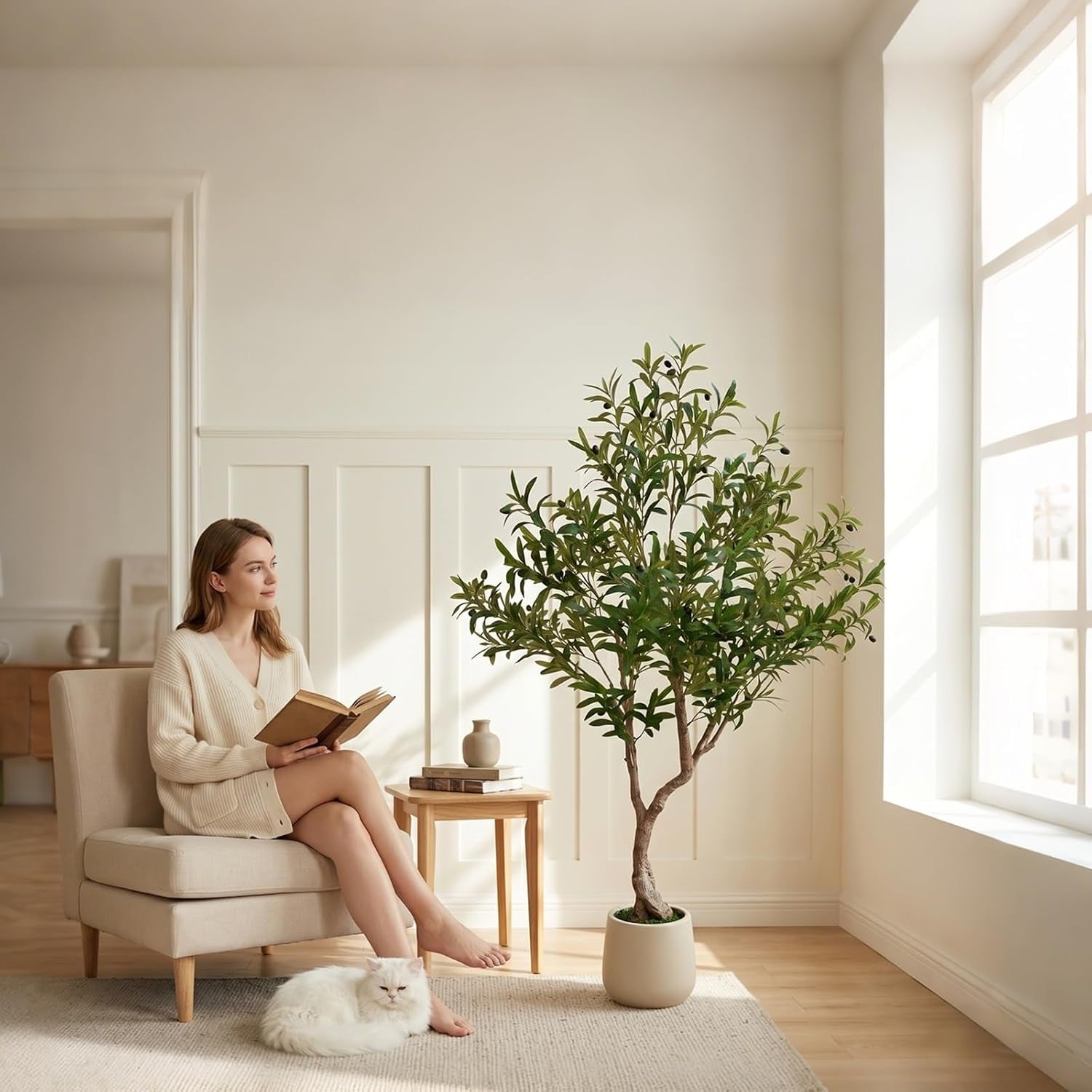 Woman reading on a beige chair next to the olive tree, cat resting on a rug in front of it.