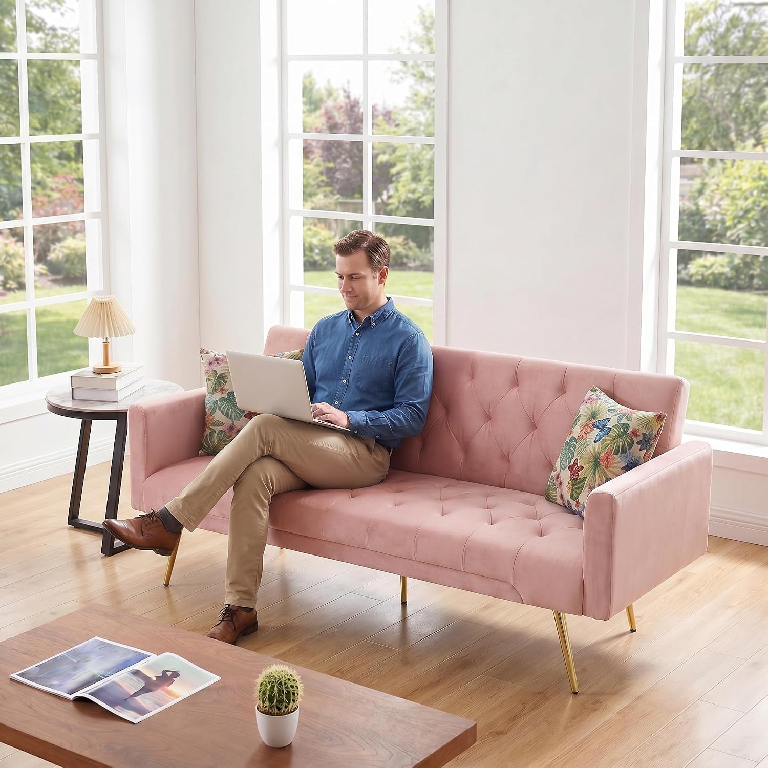 Man working on a laptop while seated on a tufted pink sofa with floral pillows beside a small round side table.